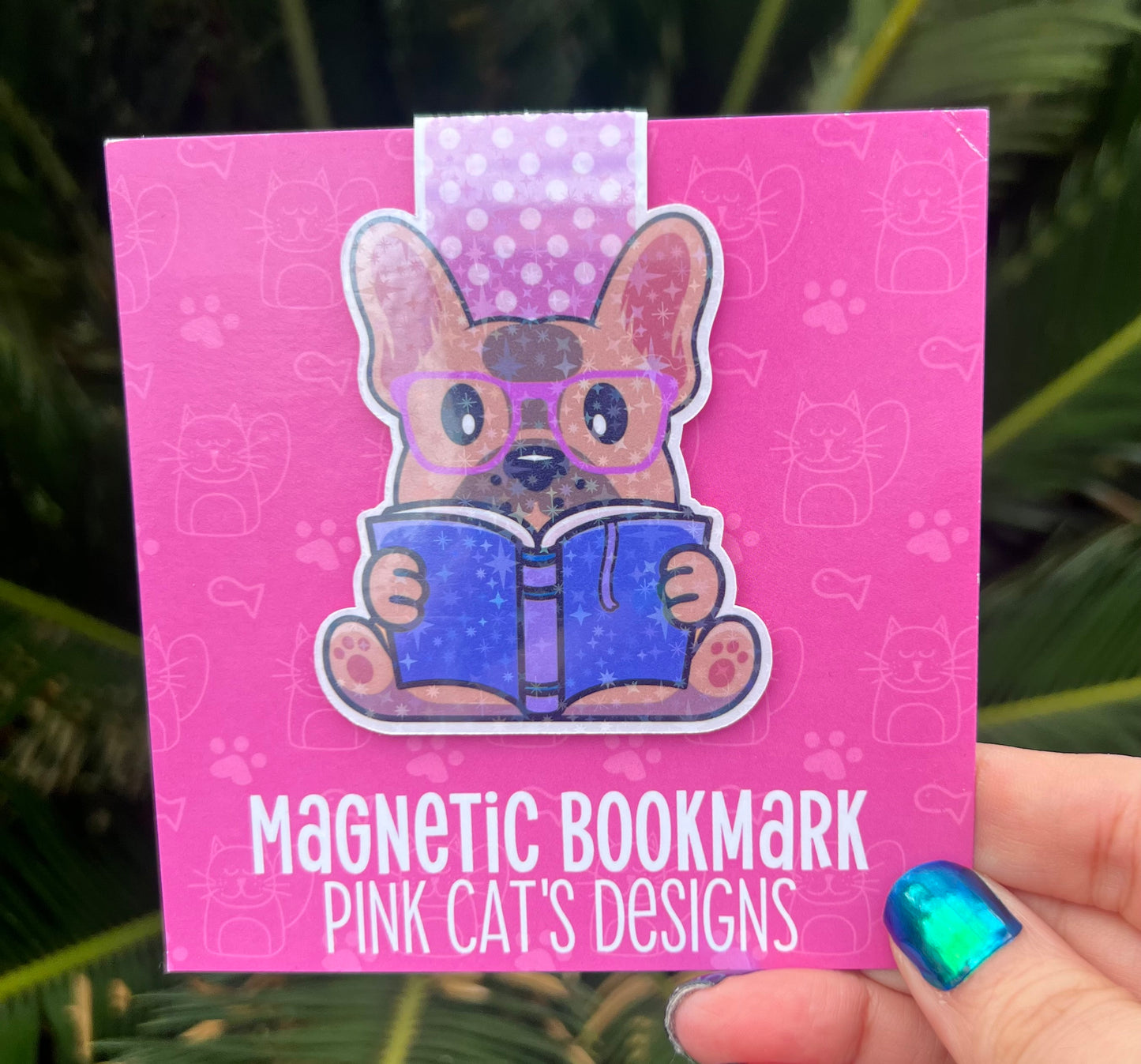 Pink magnetic bookmark with a dog design holding a book, held by a hand against a leafy background.