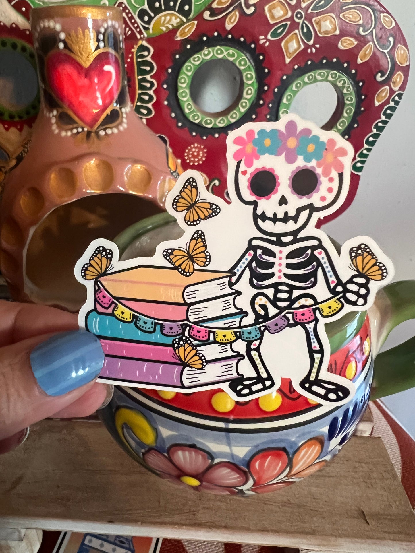 Colorful ceramic vase with a skeleton design and butterfly decorations, held by a person with blue nail polish.