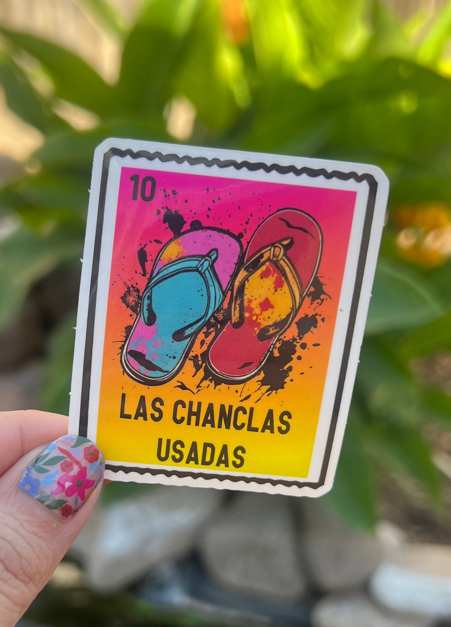 Sticker with colorful flip-flops and text 'Las Chanclas Usadas' held by a hand against a blurred green background.