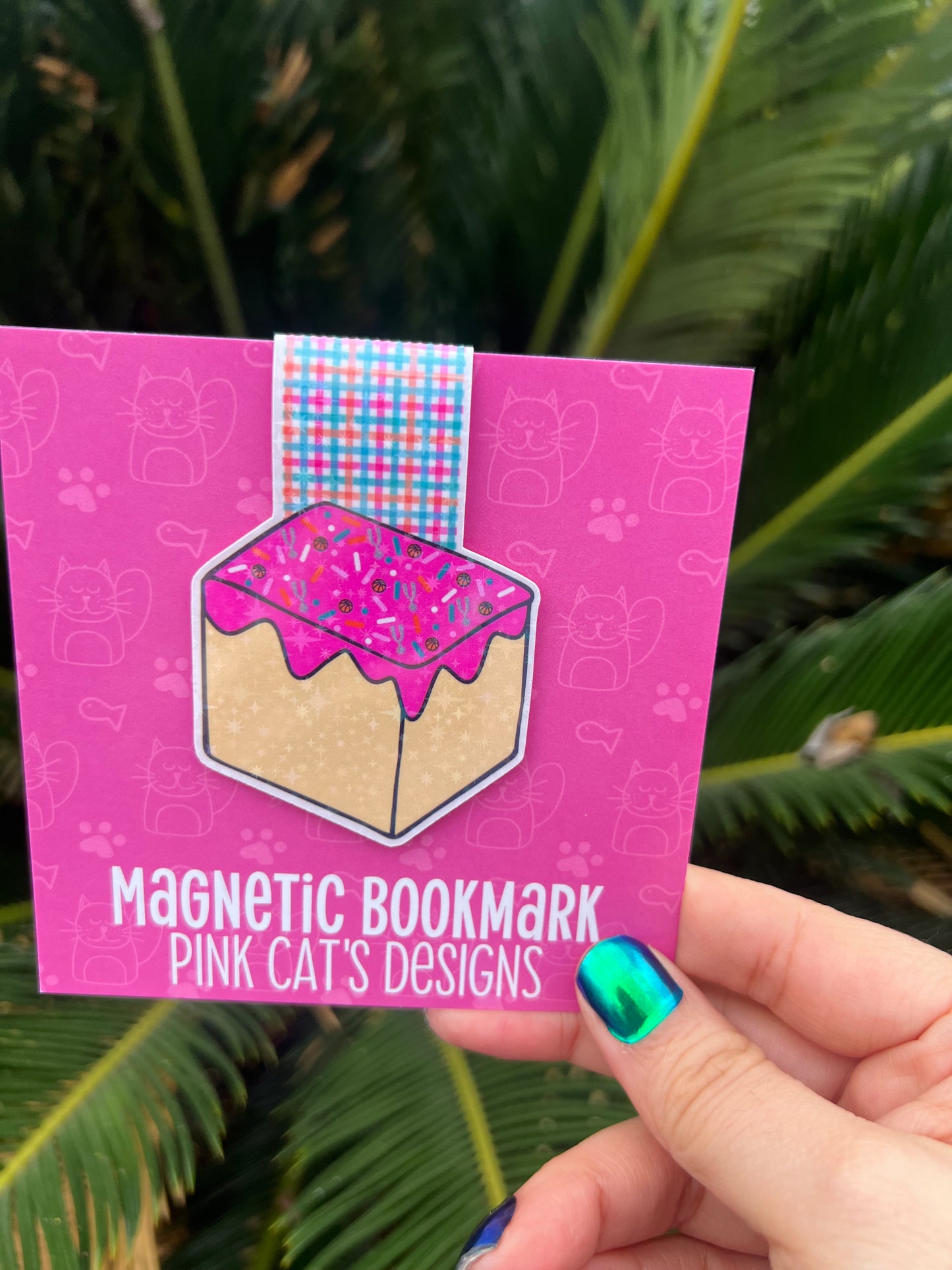 Pink magnetic bookmark packaging held by a hand with green nail polish against a green leafy background.