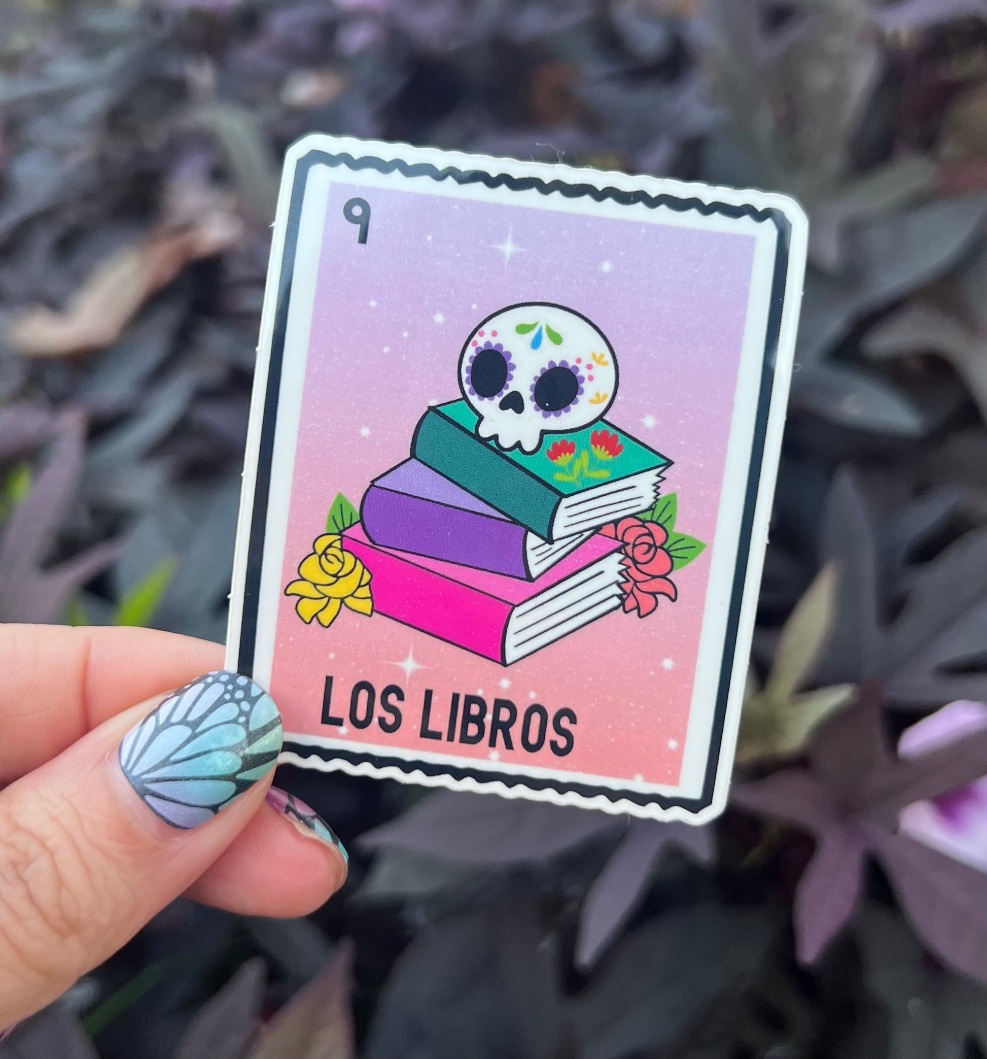 Card with a skull design and books titled 'Los Libros' held by a hand against a plant background.