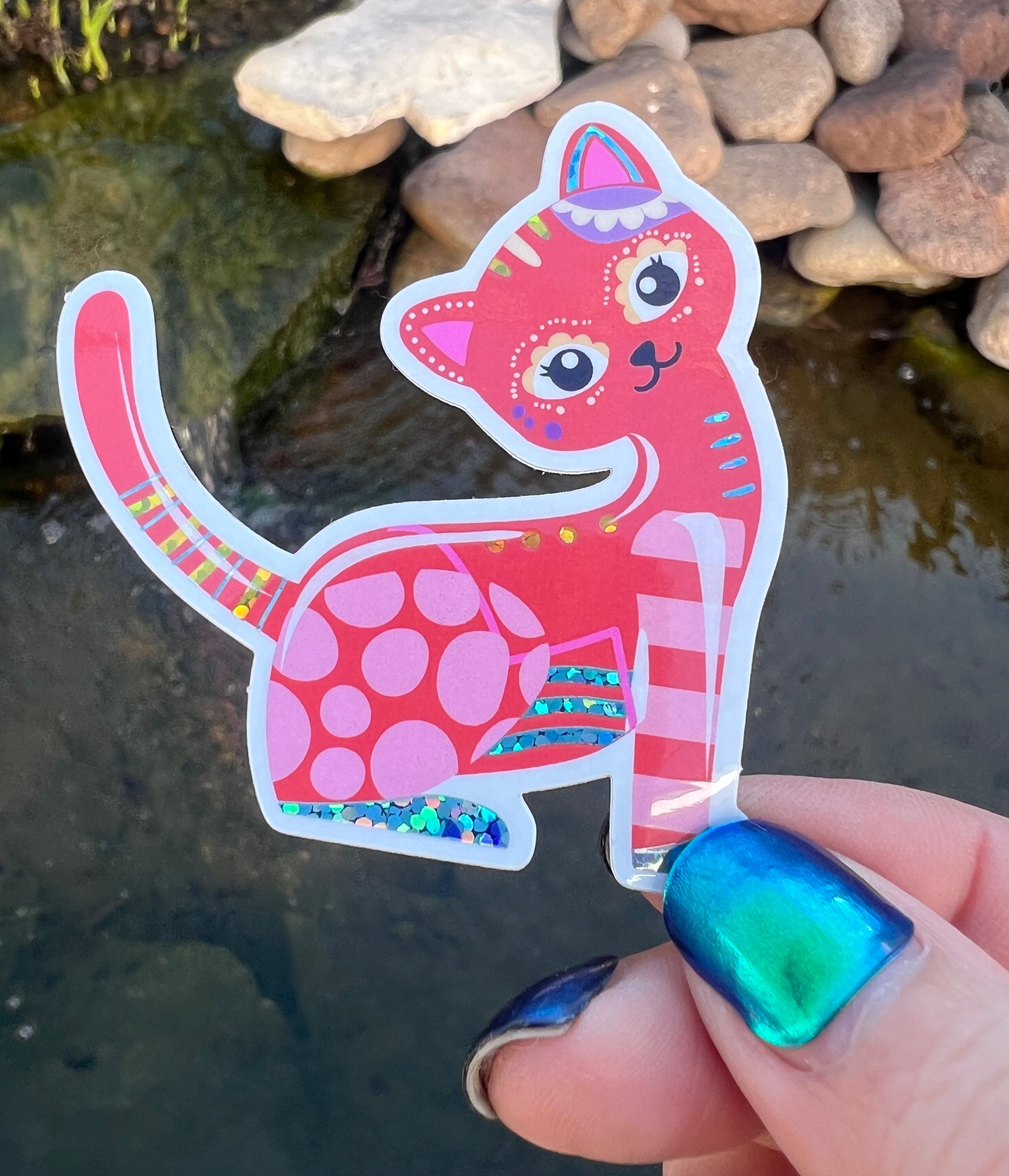 Colorful cat-shaped sticker held by a hand with blue nail polish against a natural background.
