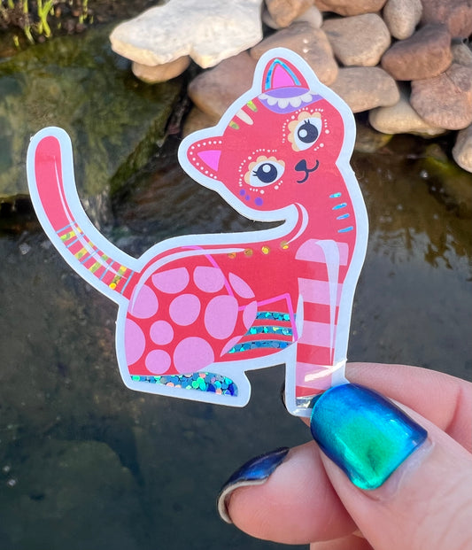 Colorful cat-shaped sticker held by a hand with blue nail polish against a natural background.