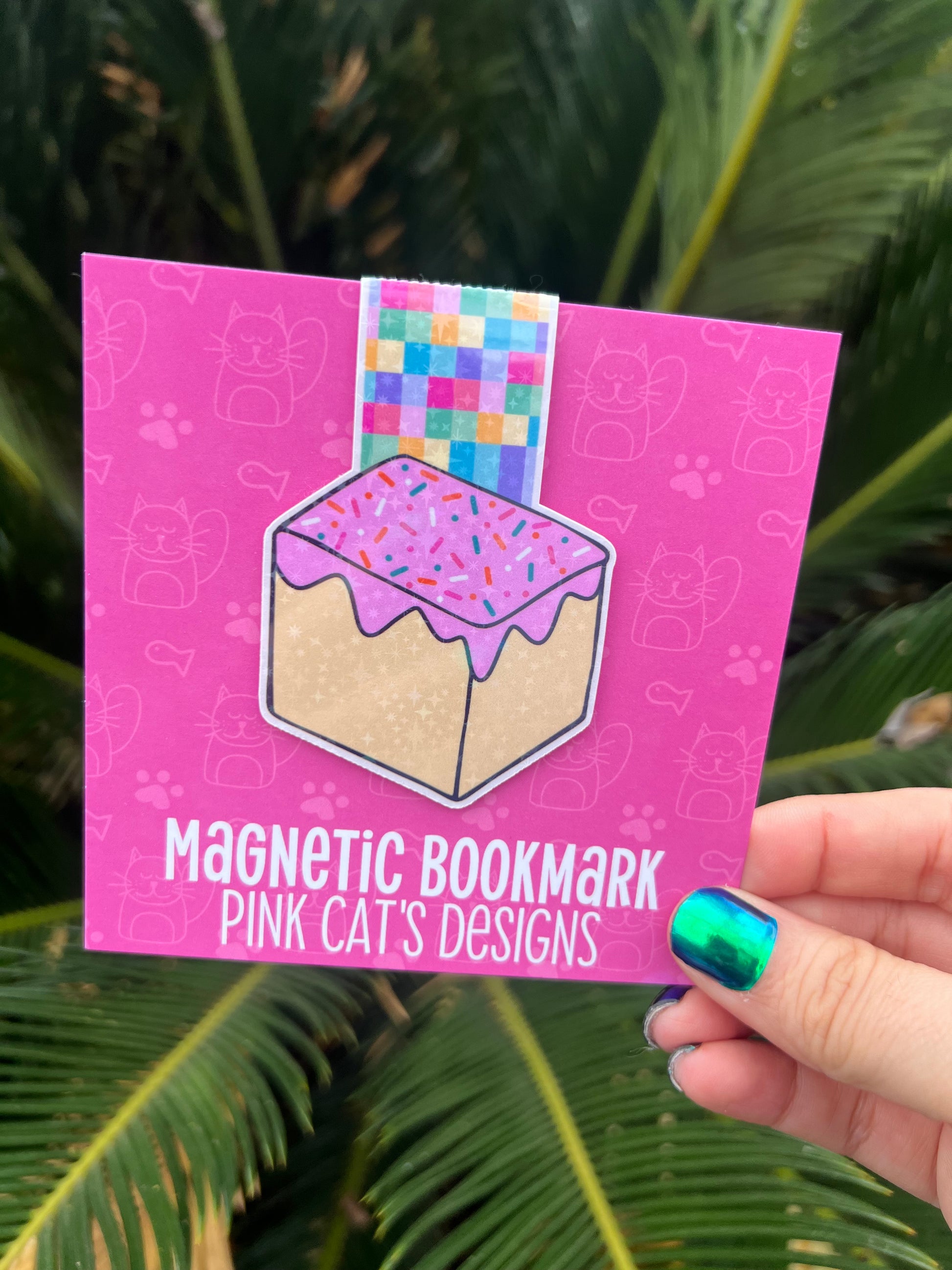 Pink magnetic bookmark with colorful design held against a green leafy background