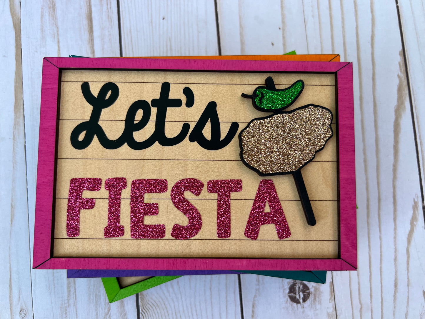 Decorative sign with 'Let's Fiesta' text and a chicken on a stick graphic on a wooden background.