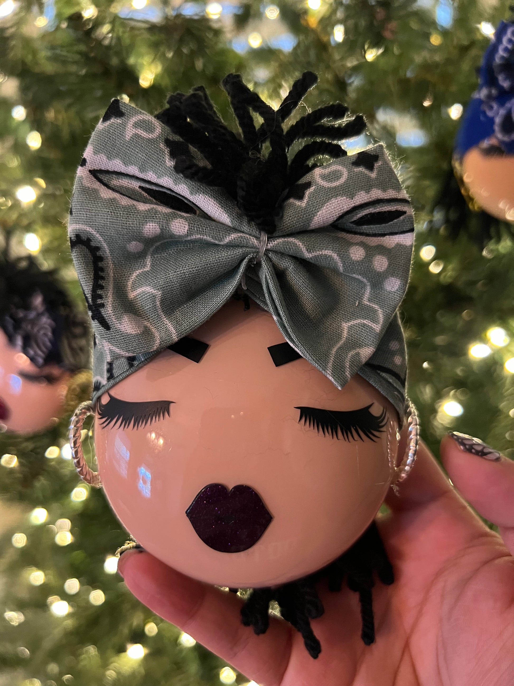 Decorative ornament with a bow and face details held in front of a Christmas tree.