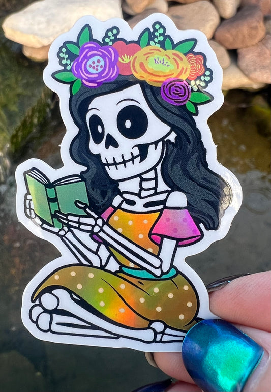 Colorful skeleton sticker with floral crown and book, held against a natural background.