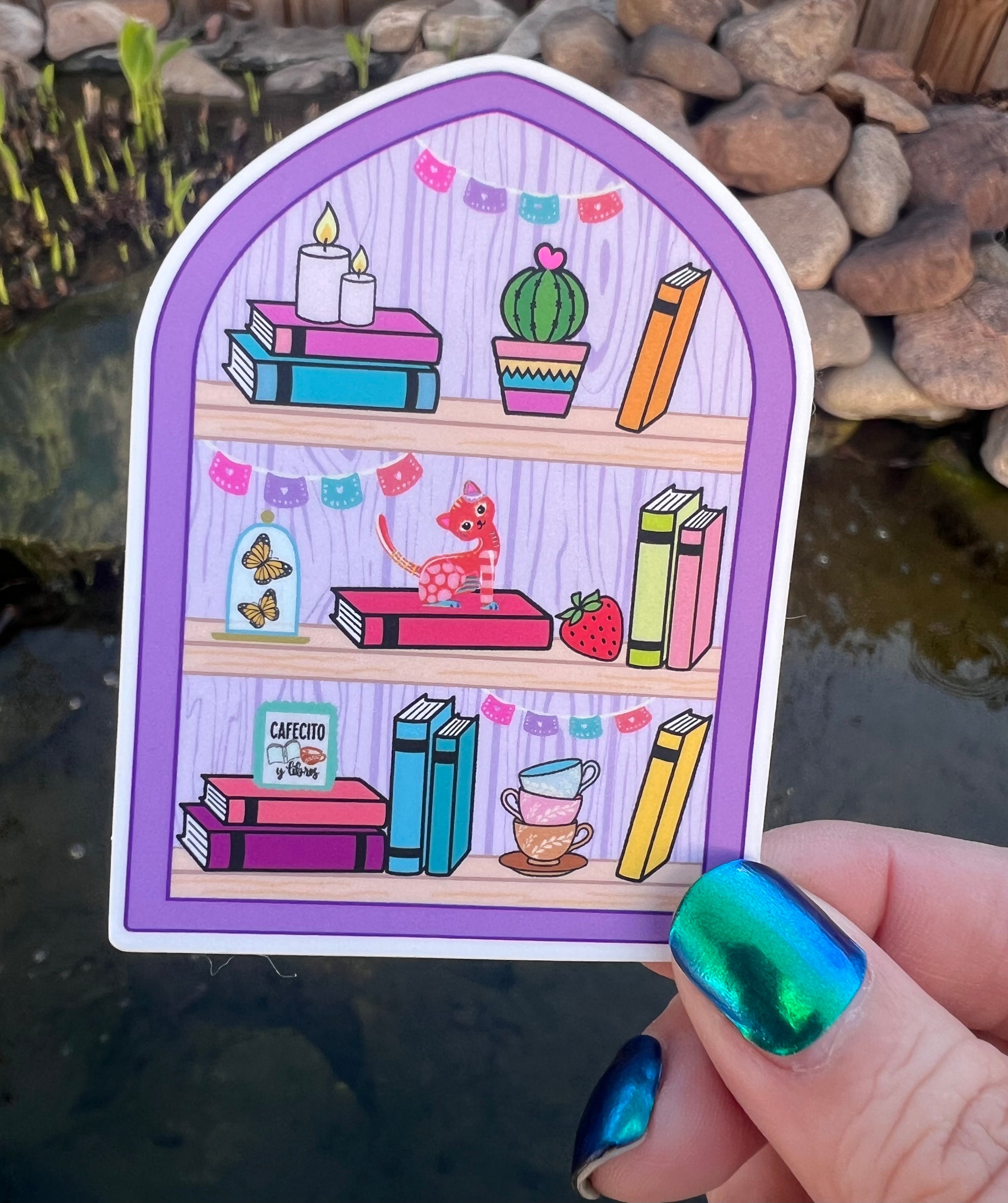 Illustrated sticker of a bookshelf scene held by a hand with colorful nail polish.