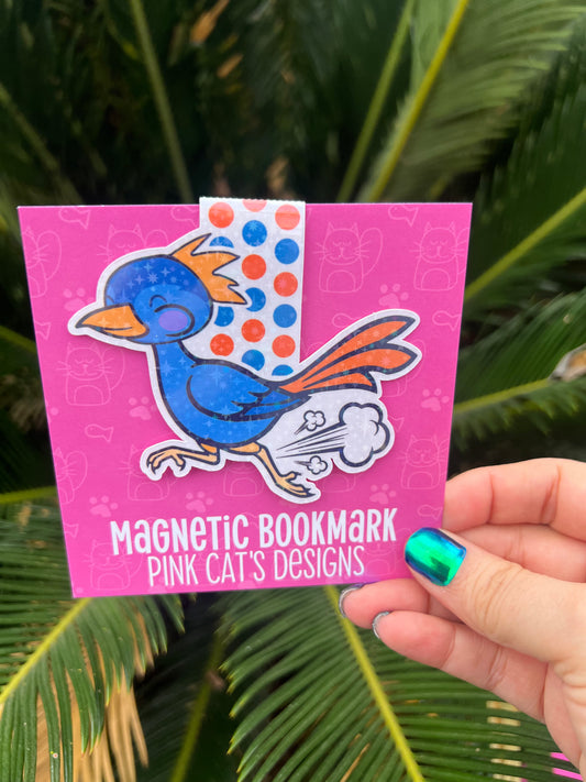 Magnetic bookmark with cartoon bird design on a pink card held by a hand against a green leafy background